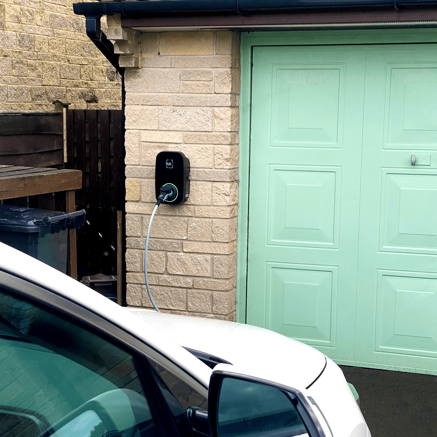 A garage with a green door and a white car in the front, with an electrical vehicle charger plugged in on a stone wall