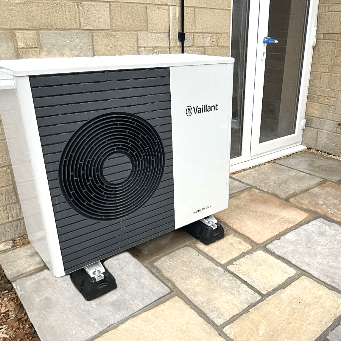An air source heat pump on a stone floor outside a house, with white doors behind it