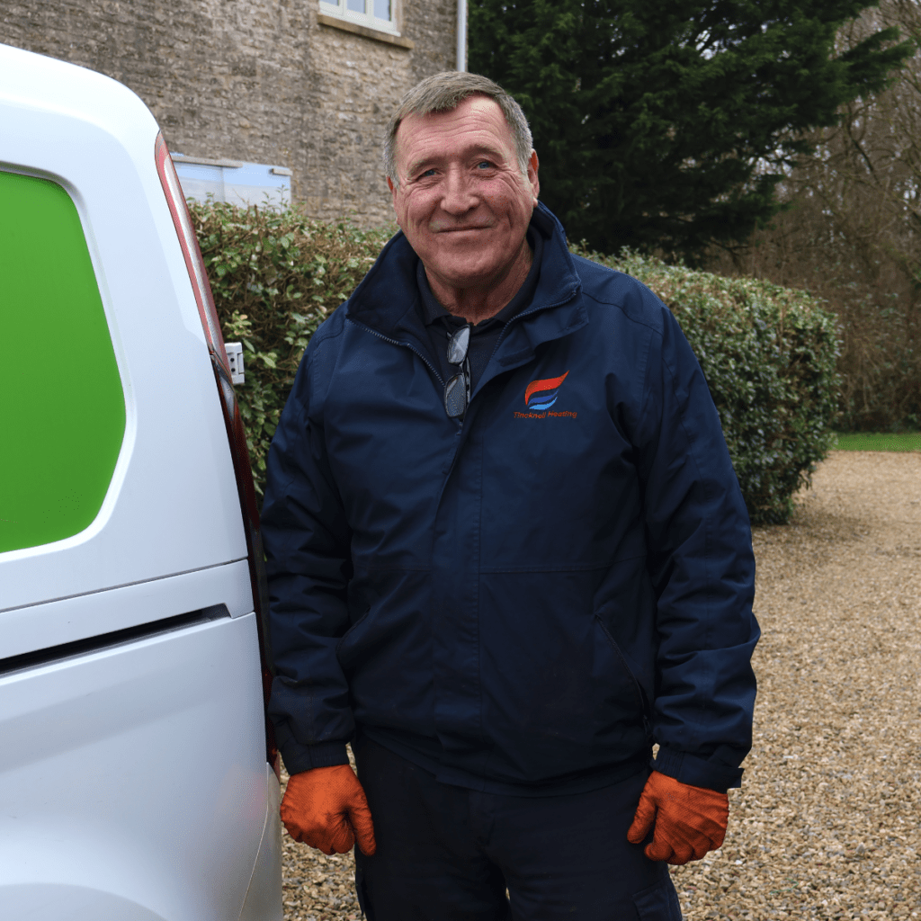 A picture of a Tincknell Heating engineer, with a blue jacket on and orange gloves. There is a white van with a green panel on it, in a garden with shingle flooring