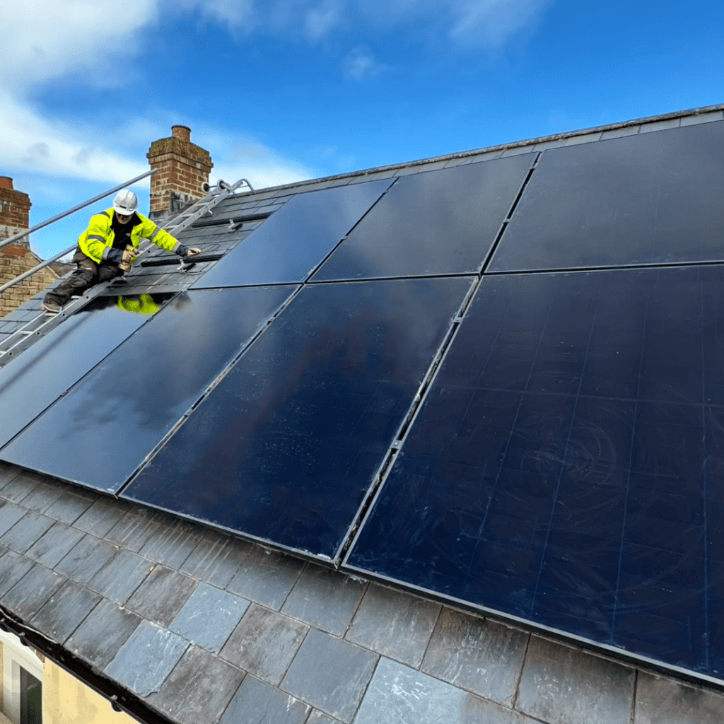 A roof with solar panels being put on. A worker in a hi-vis jacket attaching the panels. A bright blue sky. 